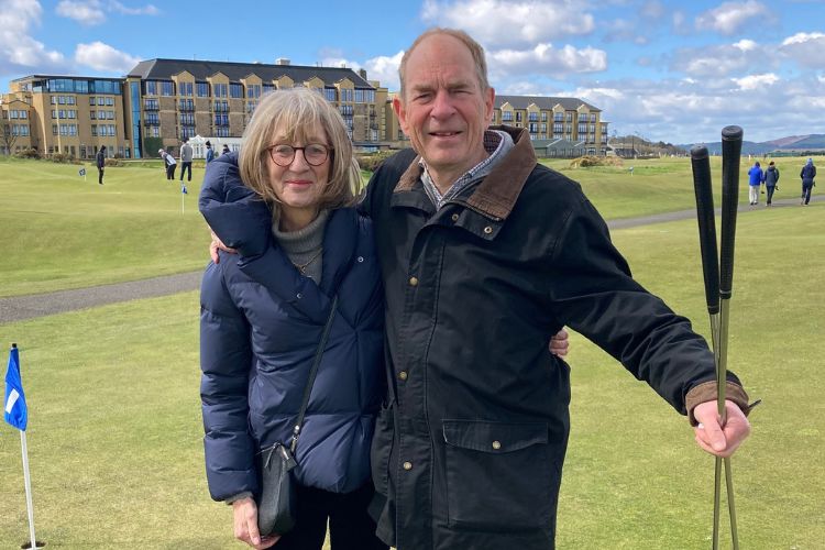 A couple, Anne and Patrick Foster, stand facing the camera. They are pictured on a golf course. Patrick is holding two putters.
