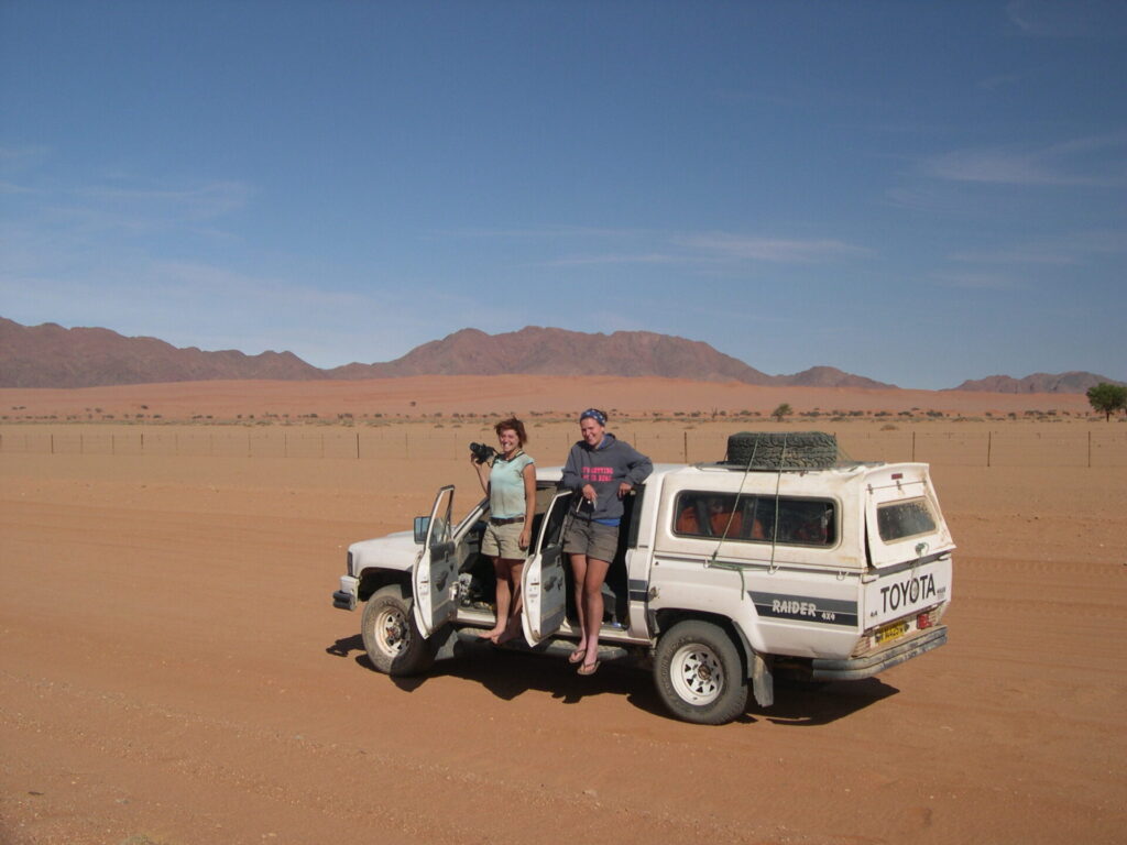 Two young women stand on the side of an off-road vehicle in a remote area of Namibia