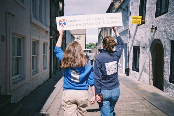 Two female students walking away from the camera on a cobbled street. Above their heads they are holding a sign advertising Geoscience courses at the University of St Andrews