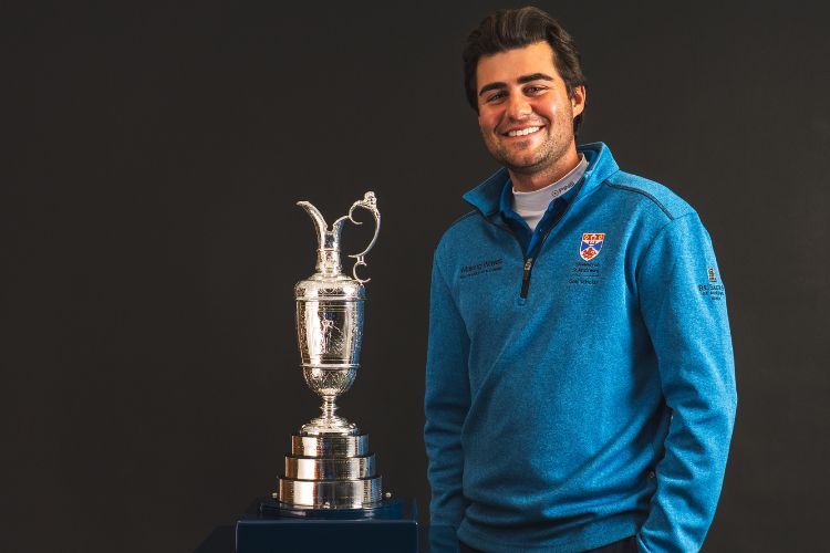 A young male, mid-twenties, poses next to the Claret Jug in a University of St Andrews branded golf top.