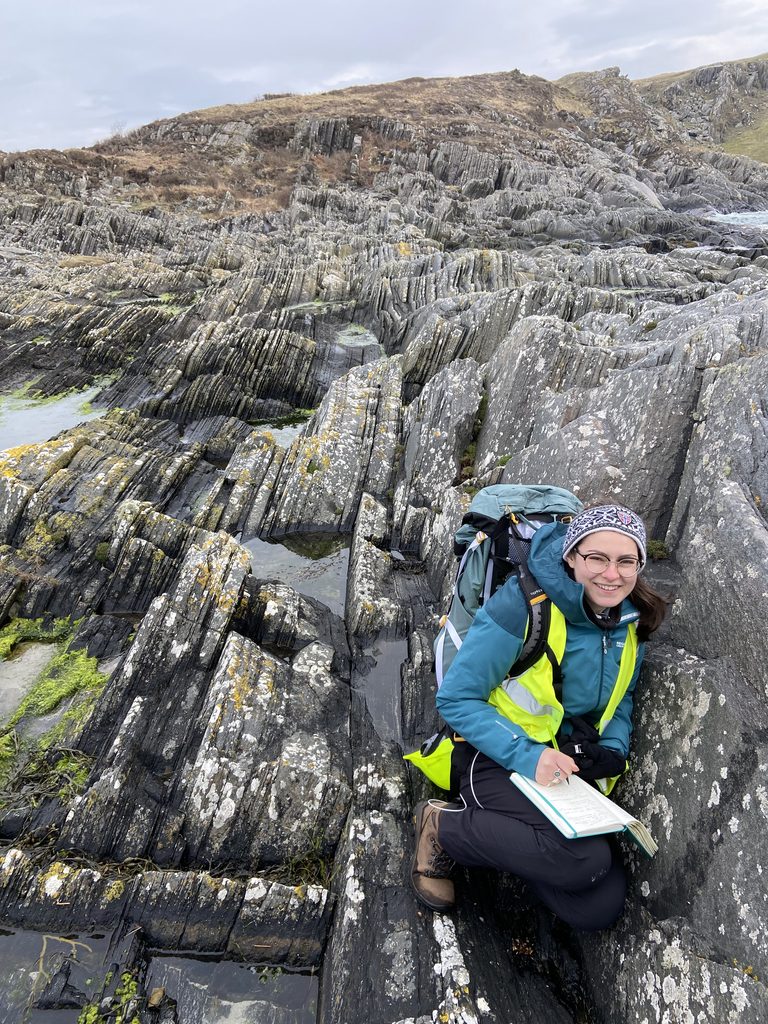 Jodie, crouching on rock formations on the Isle of Mull. She is dressed in outdoor hiking gear and is writing in a notebook