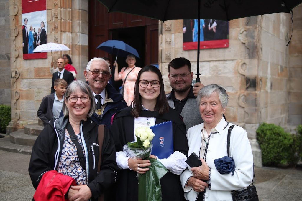 Jodie, dressed in a graduation gown and holding flowers poses for a group photo with her family in St Salvator's Quad. One member of her family holds an umbrella over the group
