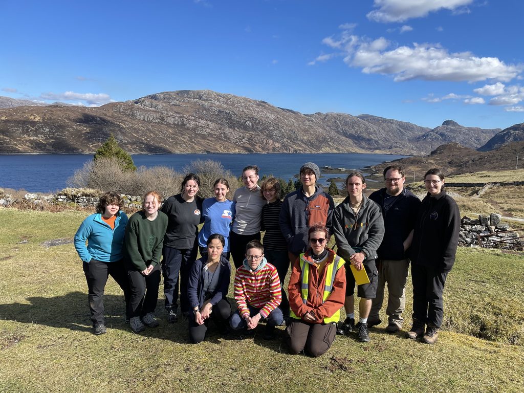 A group of 13 geology students pose for a group photo on a patch of grass. Hills and a loch can be seen in the background