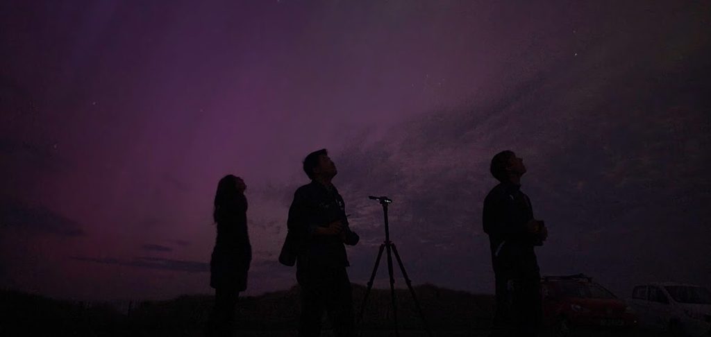 Outline of Jodie and her two friends, looking up at the nights sky illuminated in a purple colour by the Northern Lights