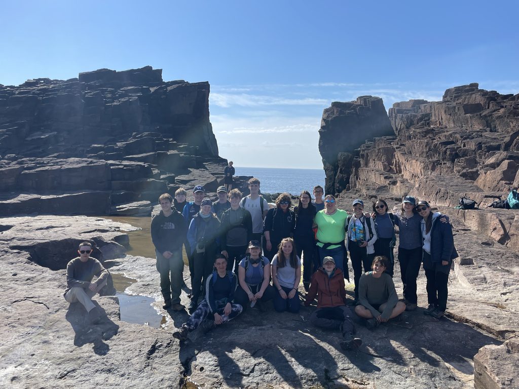 A large group of Geology Society members pose for a photo at Eshaness. Large rock formations and the sea can be seen behind the group