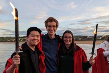 Jodie and two friends stand in red gowns holding lit torches on the Pier. The sea and St Andrews skyline can be seen in the background