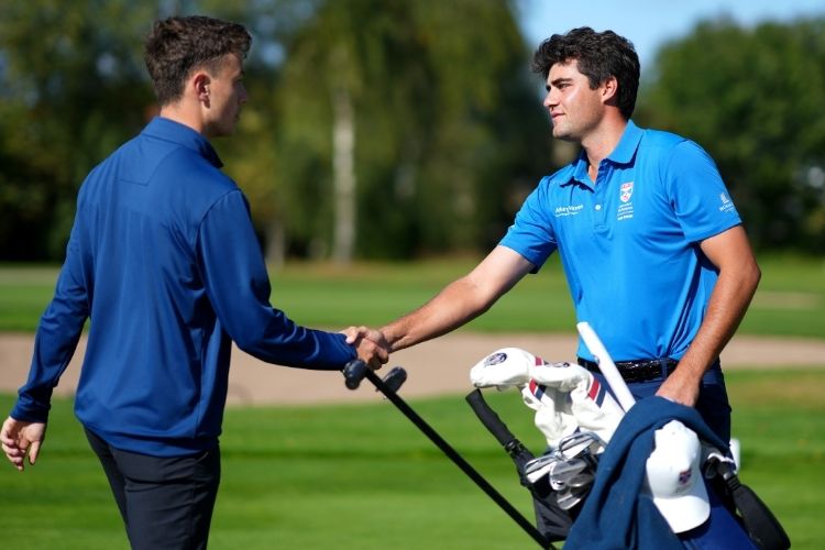 Two young male golfers shake hands on the golf course. A golf bag filled with clubs sits in the foreground.